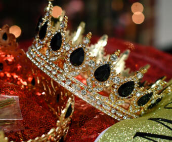 Gold tiara encrusted with clear and black gemstones rests on red sequined fabric, glittery party decor in background