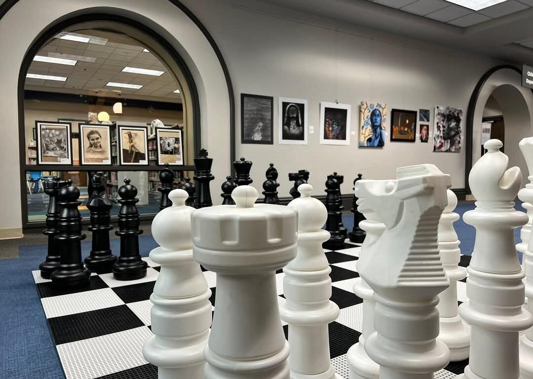Oversized white chessboard with large white chess pieces in a gallery space, black pieces visible in the background through an archway.