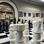 Oversized white chessboard with large white chess pieces in a gallery space, black pieces visible in the background through an archway.