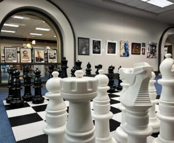 Oversized white chessboard with large white chess pieces in a gallery space, black pieces visible in the background through an archway.