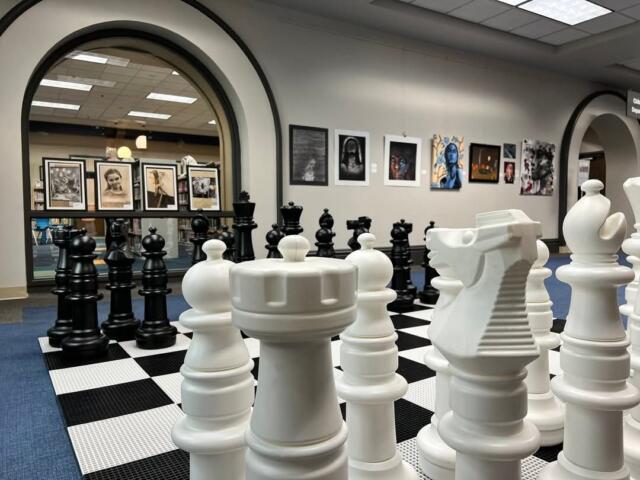 Oversized white chessboard with large white chess pieces in a gallery space, black pieces visible in the background through an archway.