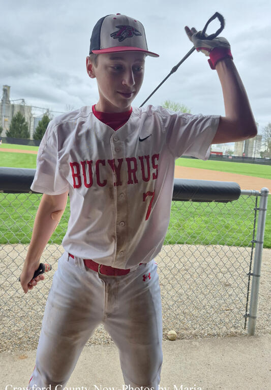 Young baseball player in a Bucyrus white jersey (#7) swings a bat near the dugout with a baseball field in the background.