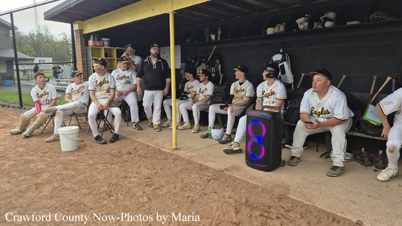 Baseball team sits in the dugout in white-and-gold uniforms, with a coach standing center and bats and gear on the wall behind.