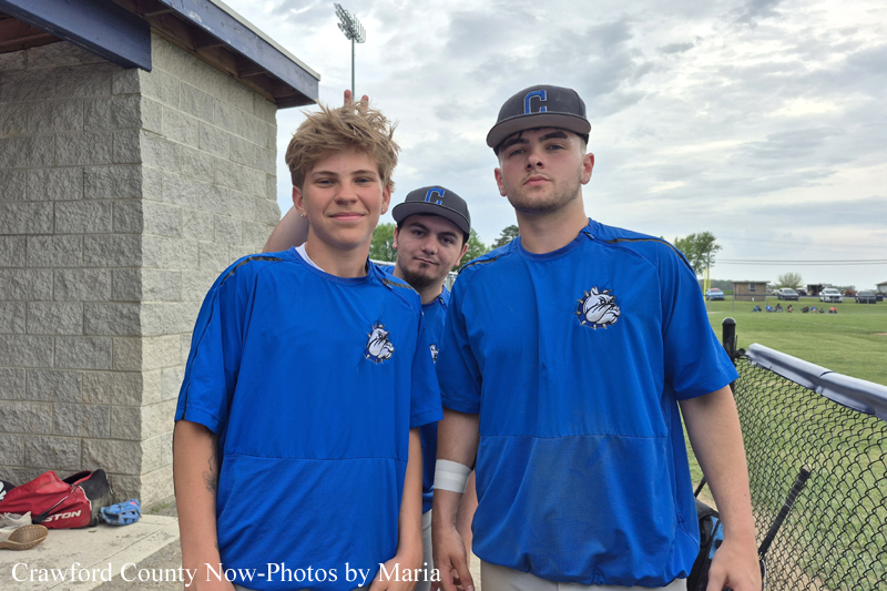 Three baseball players in blue jerseys pose together by a dugout, facing the camera with smiles.