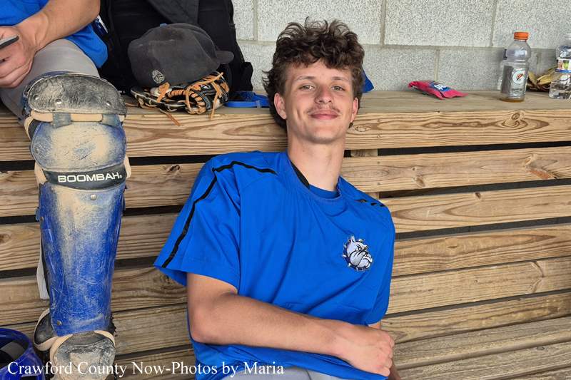Young baseball player in a blue shirt sits and smiles on a wooden dugout bench, with catcher's gear beside him.