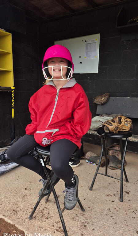 Young girl in a pink helmet and red jacket sits on a folding stool in a dugout, wearing a catcher’s facemask and baseball glove nearby, smiling at the camera.