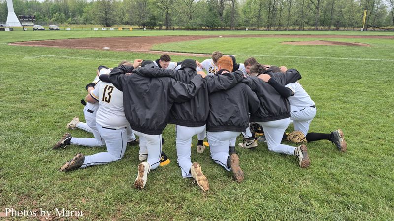 Baseball players in white uniforms form a huddle on the grass before a game on a baseball field, arms around each other in a circle.