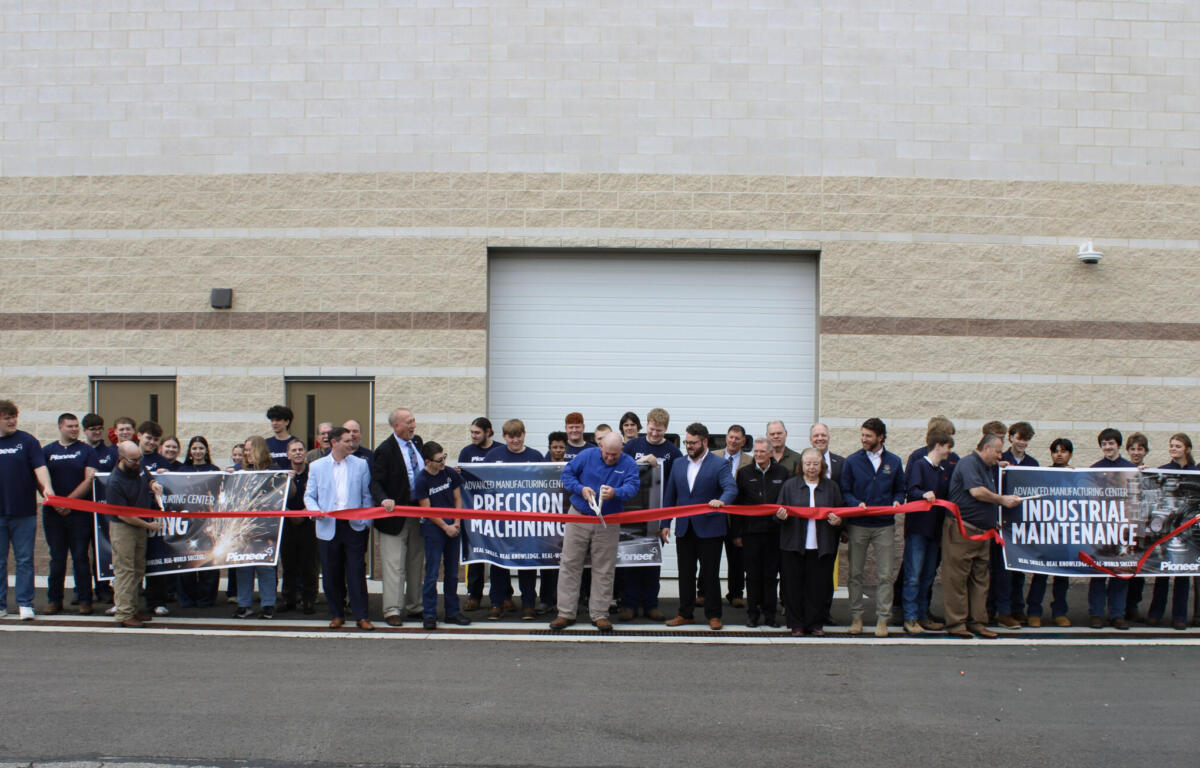 Group of people at a ribbon-cutting ceremony in front of a beige industrial building, holding banners reading industrial maintenance and precision machining.