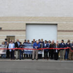 Group of people at a ribbon-cutting ceremony in front of a beige industrial building, holding banners reading industrial maintenance and precision machining.