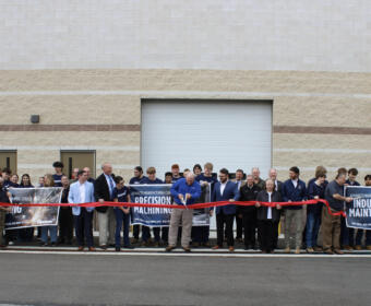 Group of people at a ribbon-cutting ceremony in front of a beige industrial building, holding banners reading industrial maintenance and precision machining.
