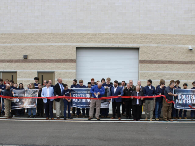 Group of people at a ribbon-cutting ceremony in front of a beige industrial building, holding banners reading industrial maintenance and precision machining.