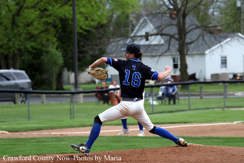 Baseball pitcher in a black jersey number 16 winds up to throw a pitch on the mound with a glove ready and blue socks in a park setting.