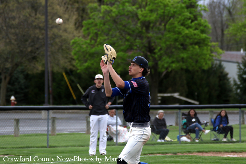 Baseball pitcher in dark uniform catches a high ball with his glove raised during a game, with spectators watching behind the fence in a grassy field.