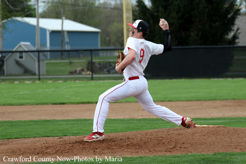 Baseball pitcher in a white uniform delivering a pitch on the mound with a green outfield behind him.