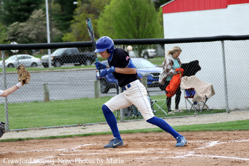 Baseball batter in blue uniform and helmet swings at a pitch on a dirt infield, with a catcher glove visible at left and spectators in the background.