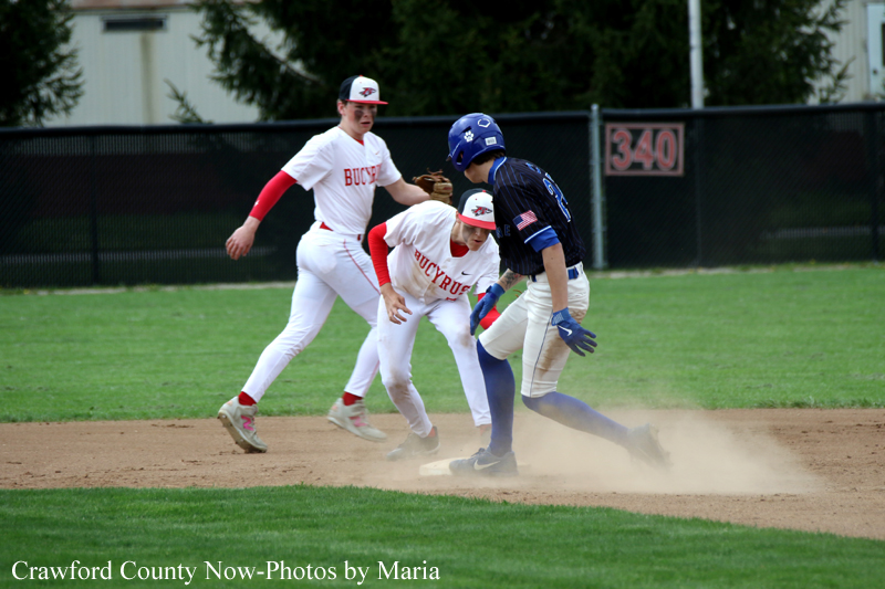 Baseball scene: a runner in blue passes a sliding into base while two players in white Buckeyes uniforms converge nearby as dust rises on the dirt infield.
