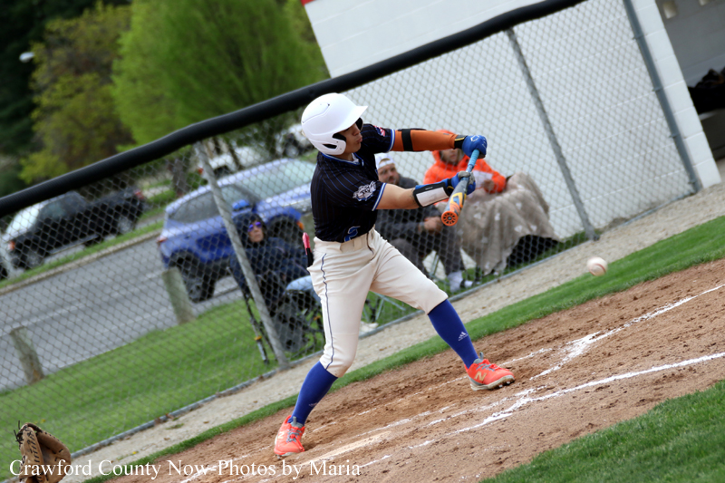 Baseball batter in white helmet and blue uniform swings at a pitch on a dirt baseball field with a fence in the background.