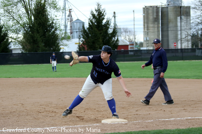 Baseball game action: a youth player in navy uniform and white pants reaches toward the ball at first base with his glove extended, while an umpire watches in the background.