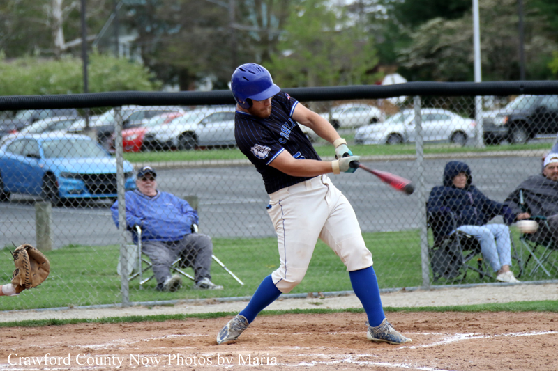 Baseball batter in a blue helmet swings at a pitch during a game, with spectators behind a chain-link fence.