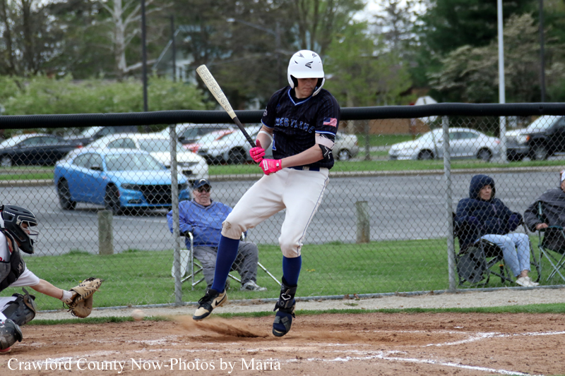 Baseball batter in a navy uniform swings at a pitch, wearing a white helmet and pink gloves at home plate with a catcher nearby and spectators in the background.
