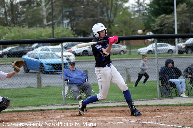 Baseball player in a navy uniform swings a bat during a game, catcher’s mitt visible on left, in a fenced field with spectators in the background.