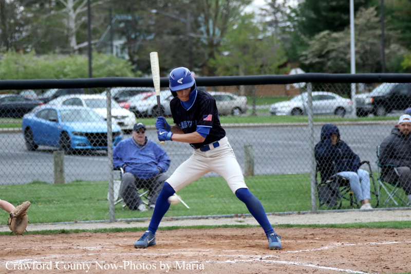 Baseball batter in a blue helmet and navy uniform stands at home plate with the bat raised, ready to swing.