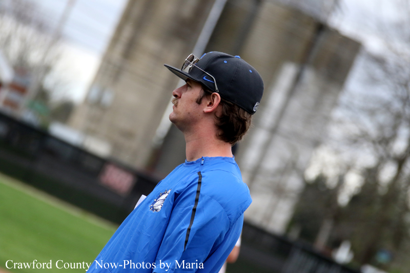 Young man in a blue sports jersey and cap stands on a baseball field, looking to the right.
