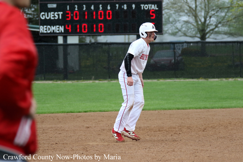 Baseball player in white uniform and helmet crouches on a dirt infield with a scoreboard behind him and a person in red in the foreground left.