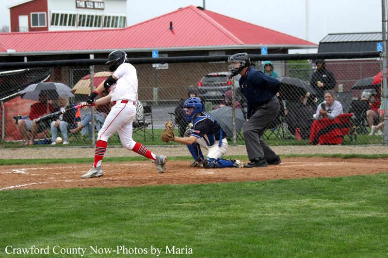 Baseball batter in white and red swings at the plate as a blue-catch catcher crouches behind, with an umpire behind them and spectators in the background.