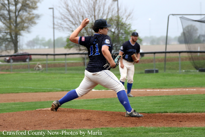 Baseball pitcher in a dark jersey throwing from the mound, with a teammate in the infield nearby on a green field overcast day.