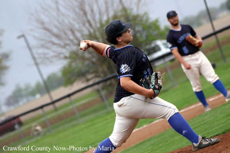 Baseball pitcher in a dark pinstripe uniform winds up to throw a pitch on the mound, a teammate watches in the background.