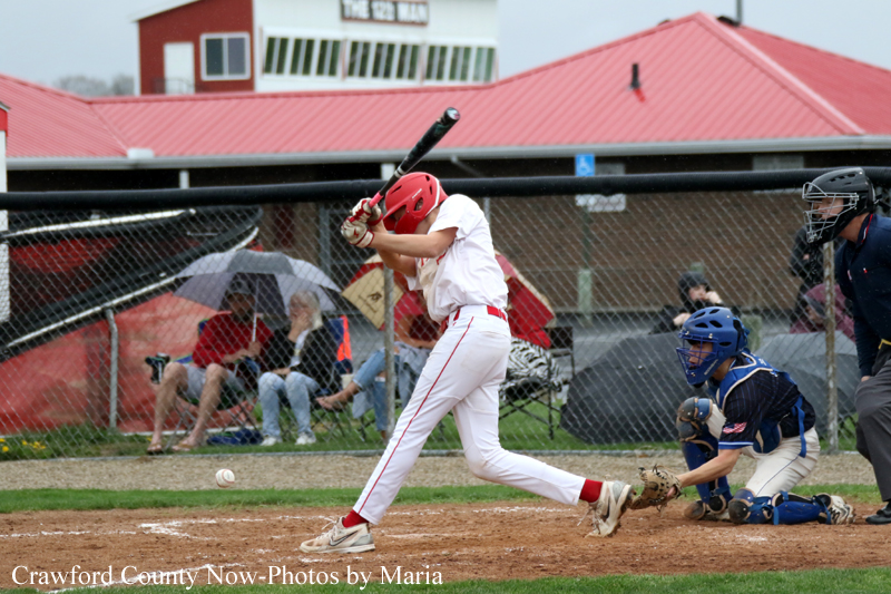 Baseball batter in a white uniform with a red helmet swings at a pitch near home plate, with a blue-catcher and umpire behind him.