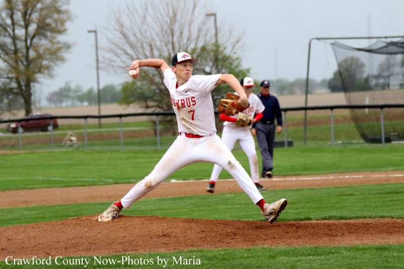 Pitcher in a white and red uniform delivers a pitch from the mound during a baseball game, with teammates and field in the background of a grassy field.