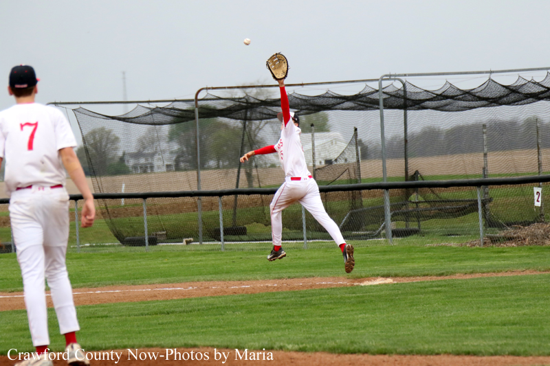 Baseball pitcher in white and red uniform mid-throw on a grassy infield, another fielder facing away nearby with netted backstop in the background.