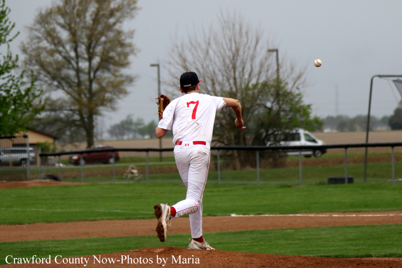 Baseball pitcher in white and red uniform (#7) throwing a pitch on the mound with a grassy field behind.