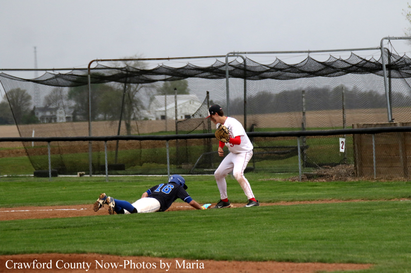 Blue-uniform baserunner slides head-first into a dirt base as a white-uniform fielder stands nearby with a glove, attempting the tag on a baseball field in action.