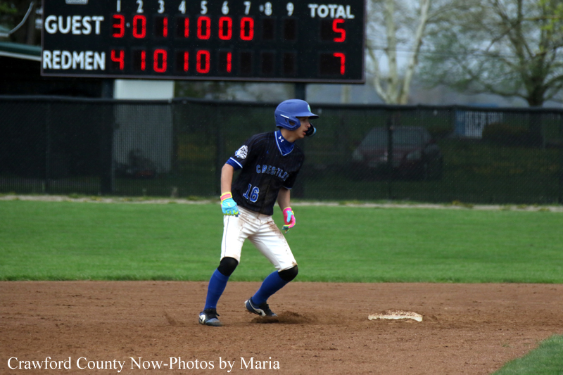 Young baseball player in a blue helmet and navy uniform stands on a base on the infield, scoreboard in the background.