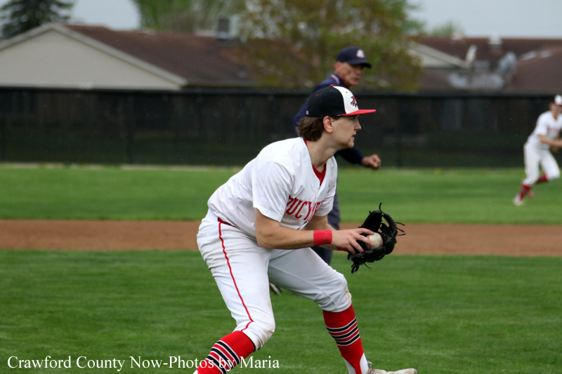 Baseball player in white and red uniform crouches with a glove, ready to field on a grassy infield.