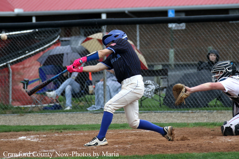 Baseball batter in a blue helmet and dark jersey swings at a pitched ball as the catcher reaches with a glove behind him on the dirt infield.