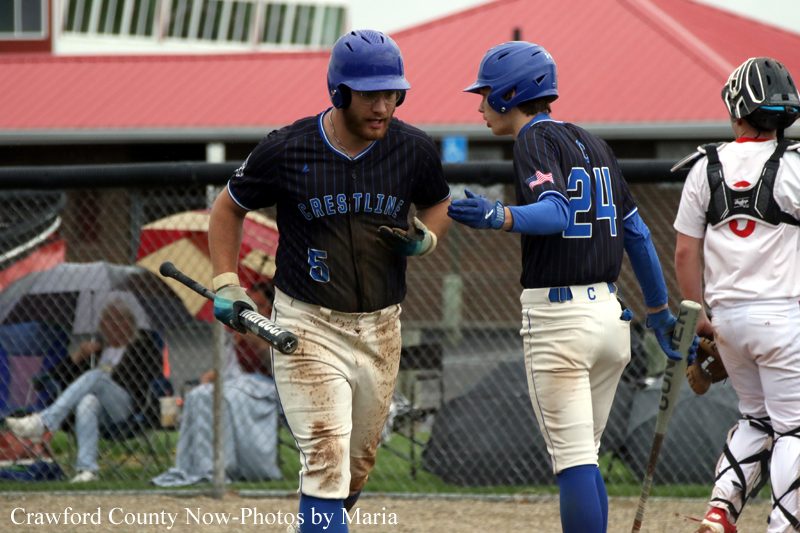 Two Crestline players wearing blue helmets share a high-five on a baseball field, one with a muddy jersey and a bat in hand, while a catcher stands nearby.