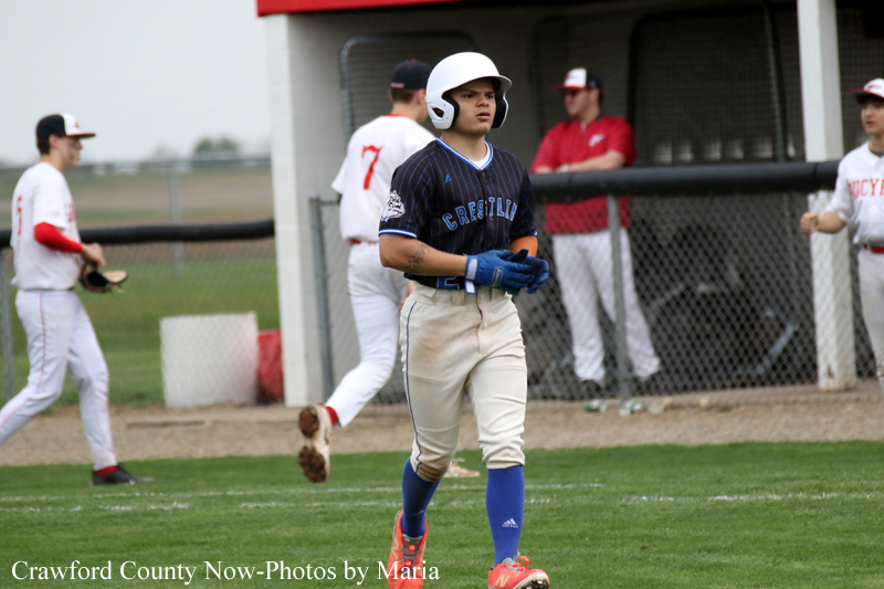 Baseball player wearing a helmet and navy uniform walks along the field near the dugout.