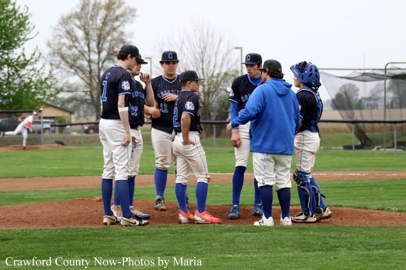 Group of young baseball players in navy uniforms listen to a coach in a blue hoodie on a baseball field around the pitcher's mound.