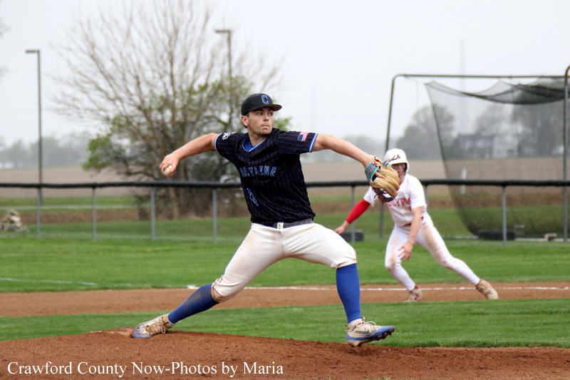 Male baseball pitcher in a dark jersey winds up to throw on the mound during a game, blue socks visible under white pants.