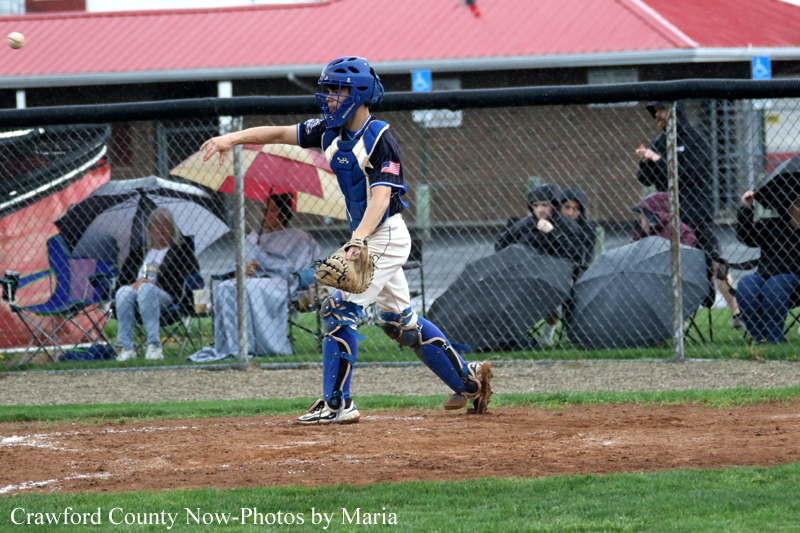 Baseball catcher in blue protective gear on the infield, glove raised, with spectators under umbrellas behind a chain-link fence.