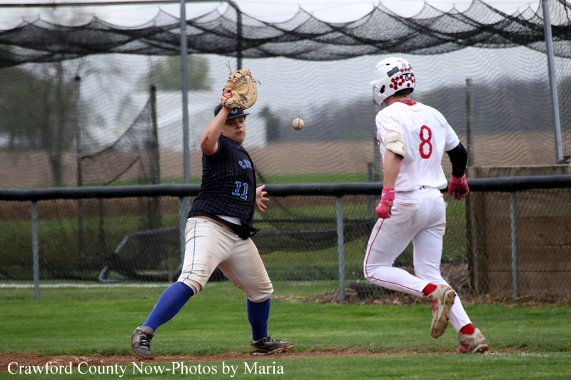 Softball pitcher in a navy uniform winds up a pitch as a runner in a white uniform (number 8) moves toward a base on a grassy field behind a chain-link fence.