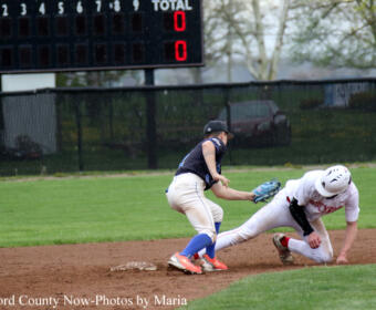 Baseball action: a runner slides into a base as a fielder in a dark uniform reaches with his glove to tag him, scoreboard in background.