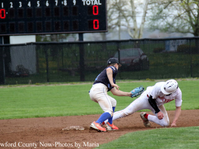 Baseball action: a runner slides into a base as a fielder in a dark uniform reaches with his glove to tag him, scoreboard in background.