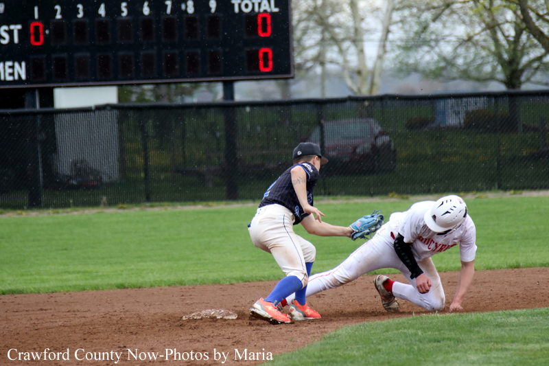 Baseball action: a runner slides into a base as a fielder in a dark uniform reaches with his glove to tag him, scoreboard in background.