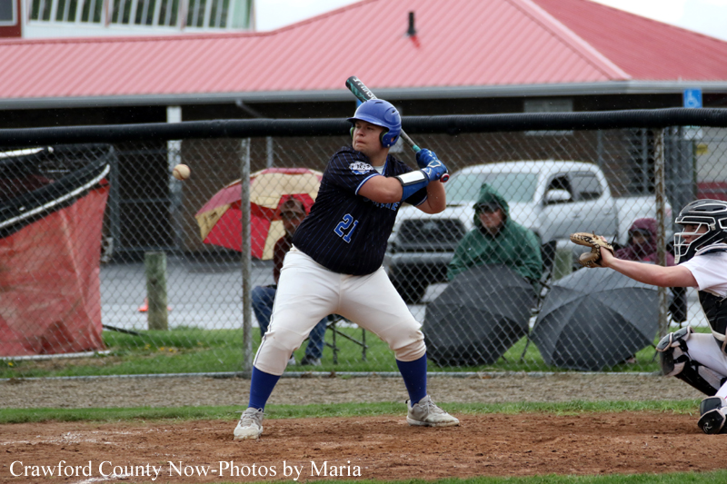 Young baseball batter in navy uniform swings at a pitch during a game, wearing a blue helmet and blue socks, with catcher and fence in the background.