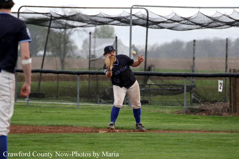 Young baseball pitcher in a navy uniform winds up to throw on the dirt mound, with a mesh backstop and outfield beyond the fence behind him.